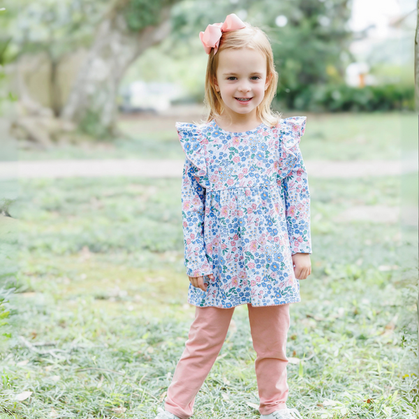 Young girl in a floral dress and pink pants standing outdoors on grass