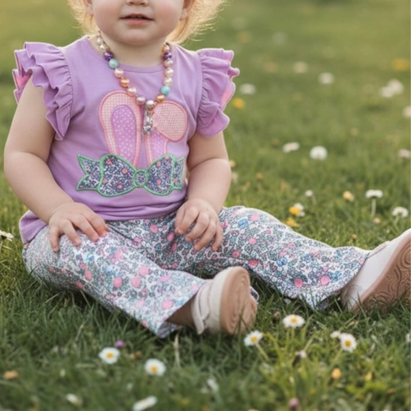 Child sitting on grass wearing a purple shirt with a bunny ear applique design and floral pants.