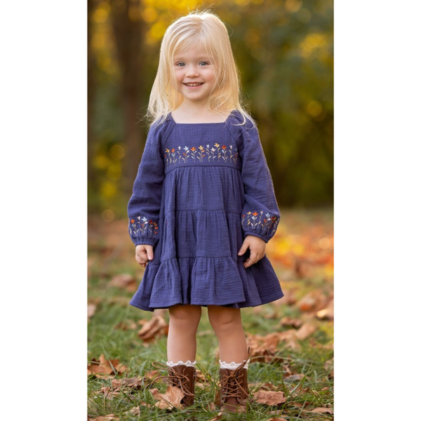 A little girl wearing a navy embroidered muslin dress, smiling outdoors in fall leaves with boots.