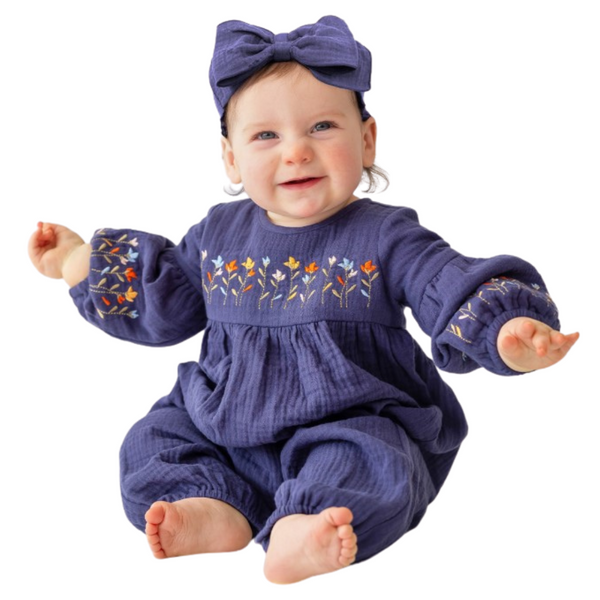 Smiling baby wearing a navy romper with embroidered flowers and puff sleeves, sitting with arms open and matching bow.