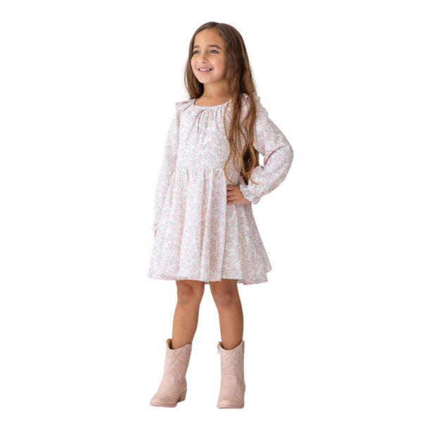 Young girl wearing a light pink floral dress with long sleeves and ruffle details, smiling with hands on hips and dressed in pink cowgirl boots.