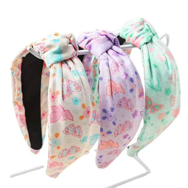A product image showing three elegant fabric headbands with a knotted design, displayed on a white wire stand. The headbands are printed with butterfly and floral patterns in pastel colors, including creamy off-white, lilac, and mint green.
