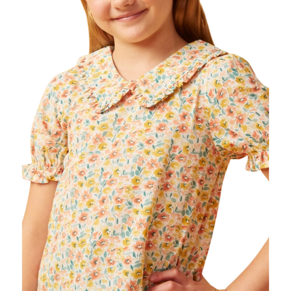 Girl wearing a floral blouse with ruffle collar and puff sleeves in coral, gold, and teal print, paired with denim jeans.