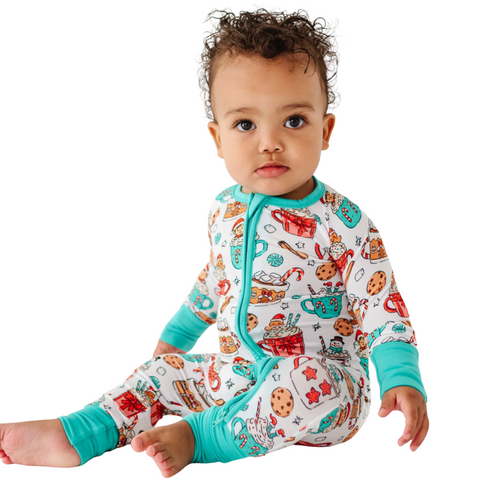A cute baby with curly hair sits on a white background, wearing a white sleeper adorned with a colorful, festive pattern of hot cocoa mugs, cookies, and candy canes, with teal cuffs and zipper trim.
