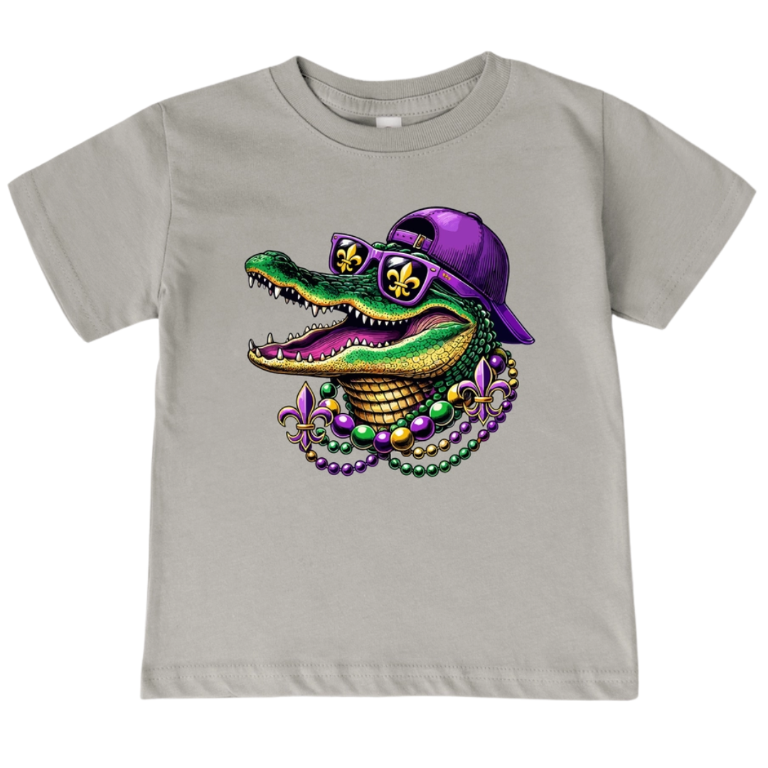 Light grey kids' t-shirt with a colorful graphic of an alligator wearing a purple backwards baseball cap, yellow sunglasses with fleur-de-lis, and purple, green, and gold Mardi Gras beads.