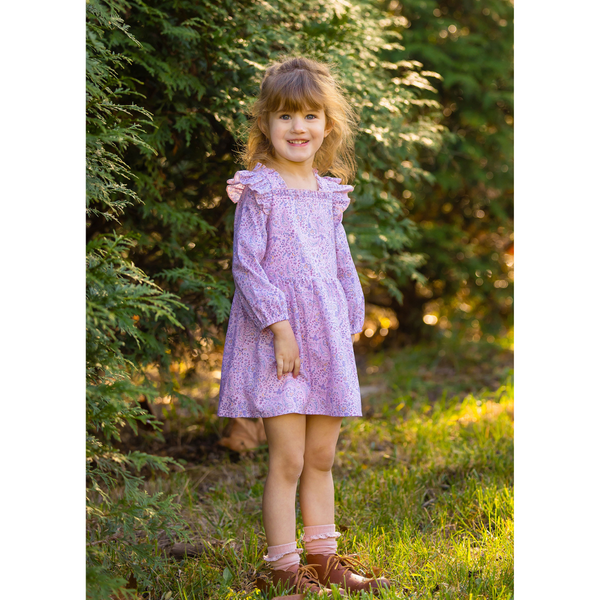 Young girl standing outdoors wearing a pink paisley print dress with long sleeves and ruffle trim, smiling in front of green foliage.

