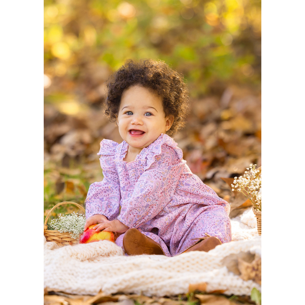 Smiling baby girl sitting on a cream knit blanket in a pink paisley romper with ruffle sleeves, surrounded by fallen leaves and warm light.