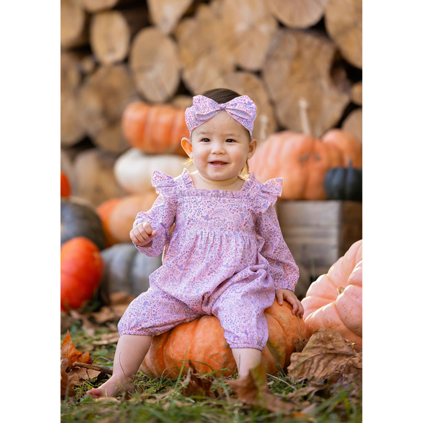 Baby girl wearing a matching paisley romper and headband, sitting on a pumpkin in front of stacked firewood and autumn gourds.

