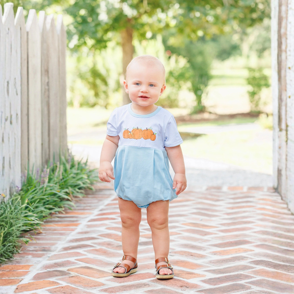 A toddler standing on a brick path outdoors, wearing a light blue romper dress with a pumpkin graphic on the chest and brown sandals.
