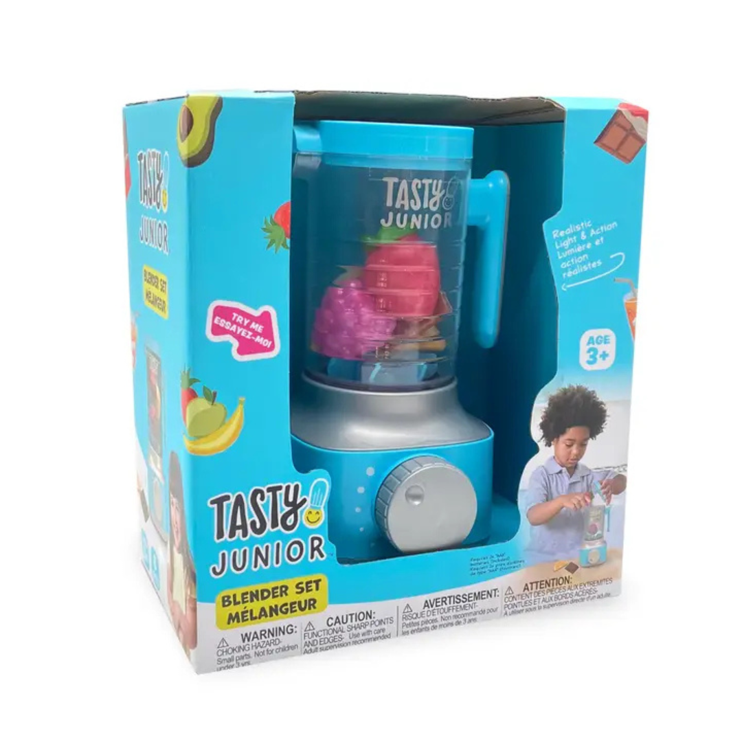 A young boy with blonde hair is sitting at a wooden table, playing with a light blue and silver toy blender from the Tasty Junior brand. He is putting a toy raspberry into the clear blender cup, which already contains other toy fruit.

