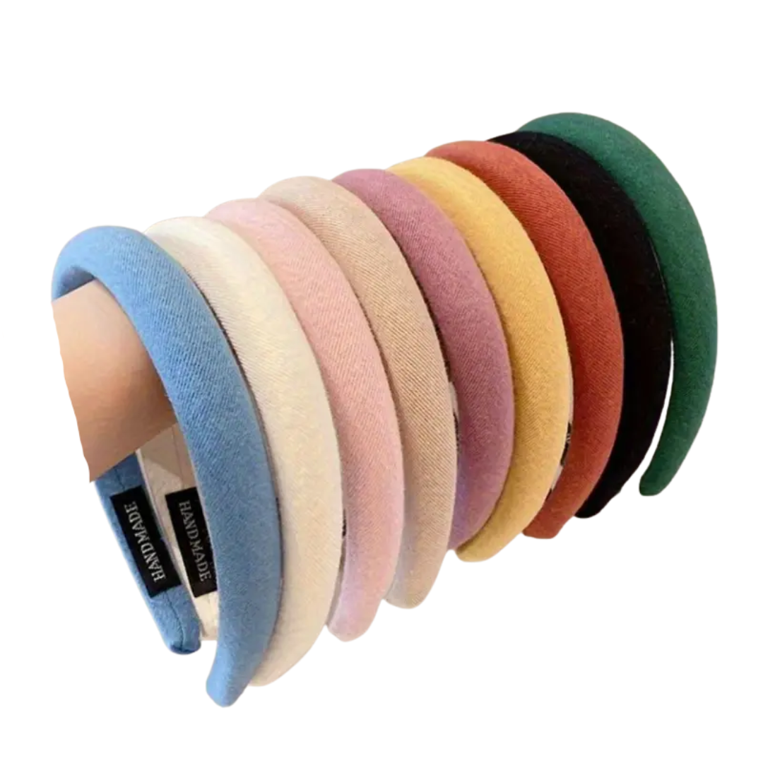 A row of eight colorful padded headbands, held by a hand, showcasing shades of light blue, ivory, soft pink, beige, light purple, yellow, rust orange, and black, each with a "HANDMADE" tag.
