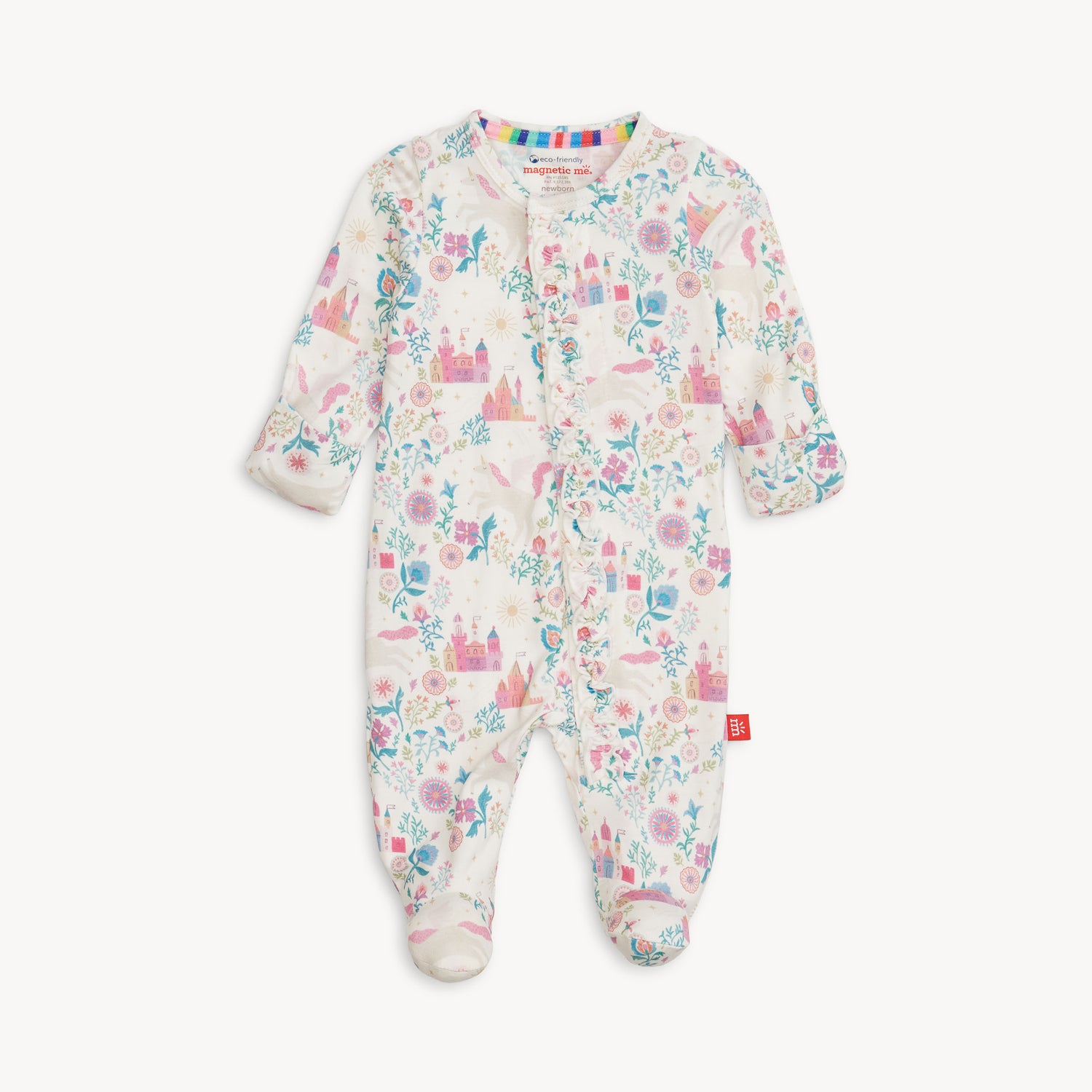 A full-length view of a white baby footie pajama with a whimsical "Tail of Magic" pattern. The pattern features colorful castles, unicorns, flowers, and leaves in shades of pink, blue, green, and purple. The one-piece pajama has long sleeves, covered feet, and a full front opening with magnetic closures. A small rainbow stripe is visible on the inner collar. The brand name, "magnetic me," is visible on a small tag. A small red logo tag is also sewn onto the lower right side.
