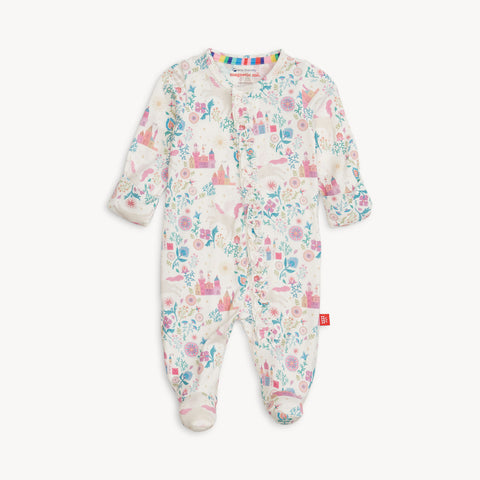 A full-length view of a white baby footie pajama with a whimsical "Tail of Magic" pattern. The pattern features colorful castles, unicorns, flowers, and leaves in shades of pink, blue, green, and purple. The one-piece pajama has long sleeves, covered feet, and a full front opening with magnetic closures. A small rainbow stripe is visible on the inner collar. The brand name, "magnetic me," is visible on a small tag. A small red logo tag is also sewn onto the lower right side.