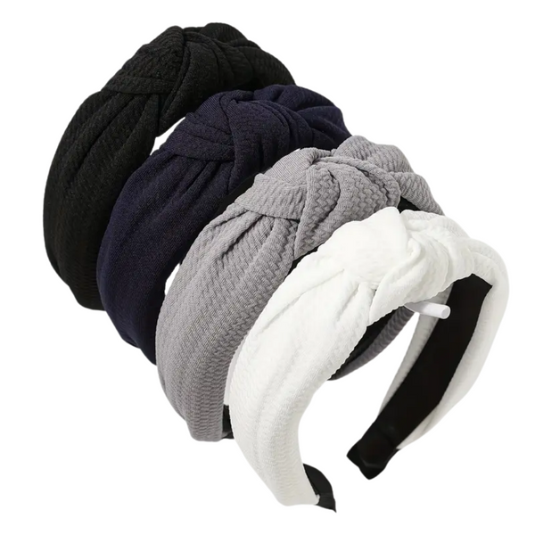 A product image showing four wide, knotted headbands stacked and fanned out. The headbands are made of a waffle-textured fabric in solid neutral colors: black, navy, gray, and white.